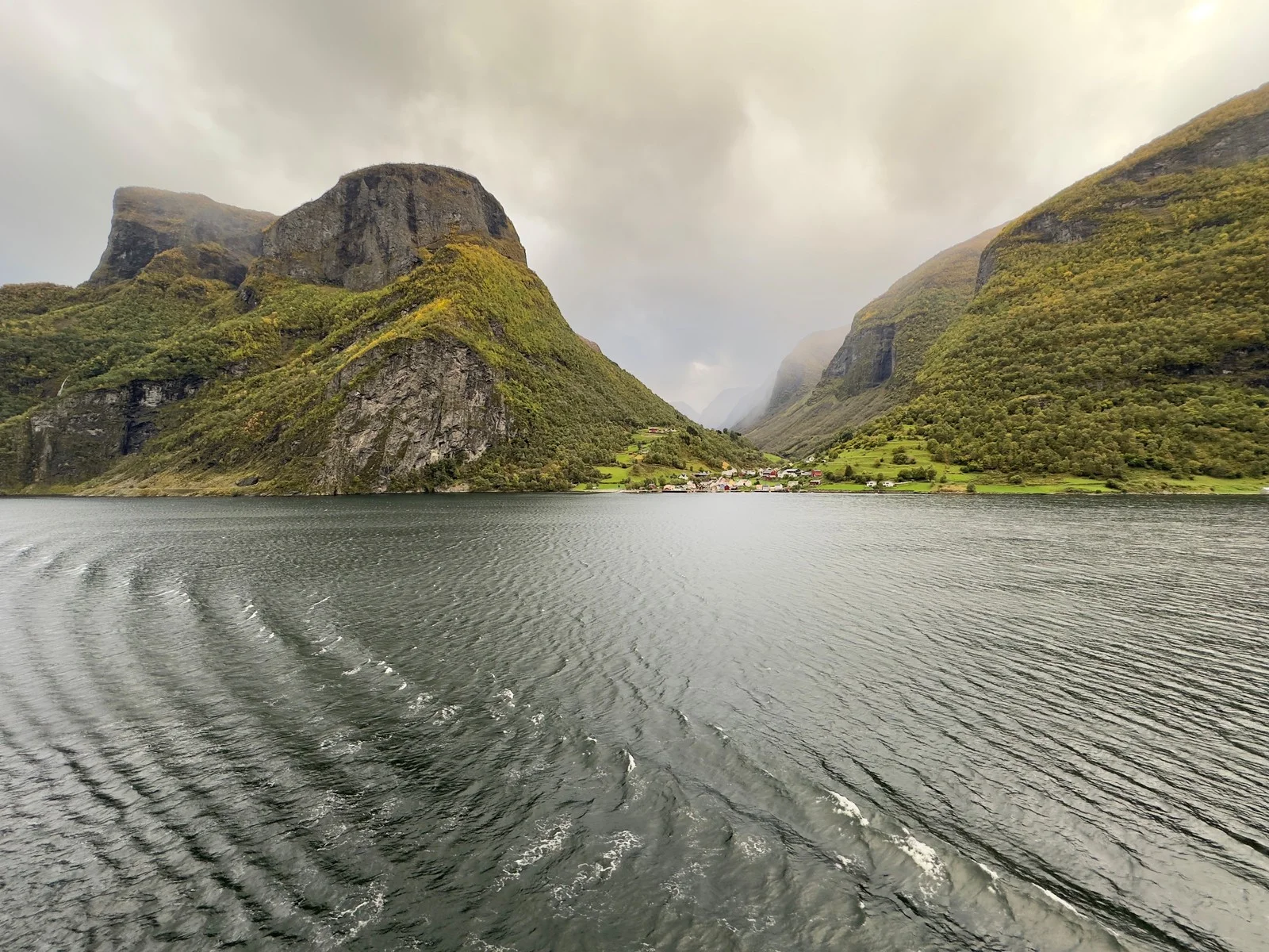 Fiordo tra montagne sotto un cielo coperto, scia dell'acqua in primo piano.